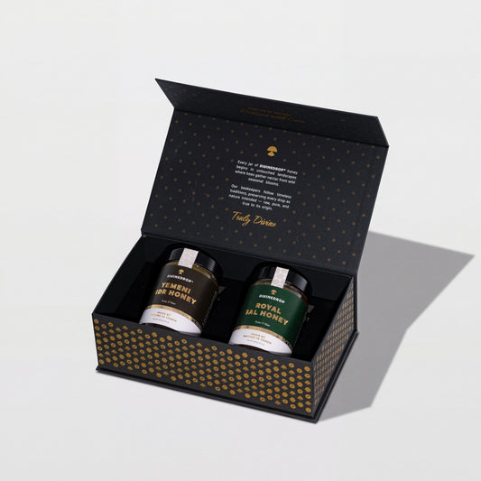 DIVINEDROP black gift box with gold pattern, open, holding two jars of Yemeni honey, on a light grey background