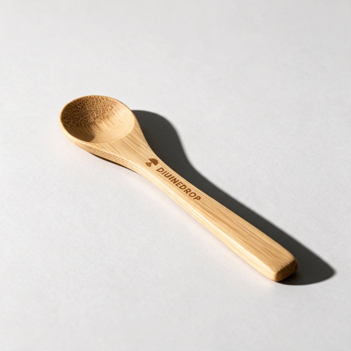Bamboo honey spoon with the DIVINEDROP logo engraved on the handle, lying on a light grey background