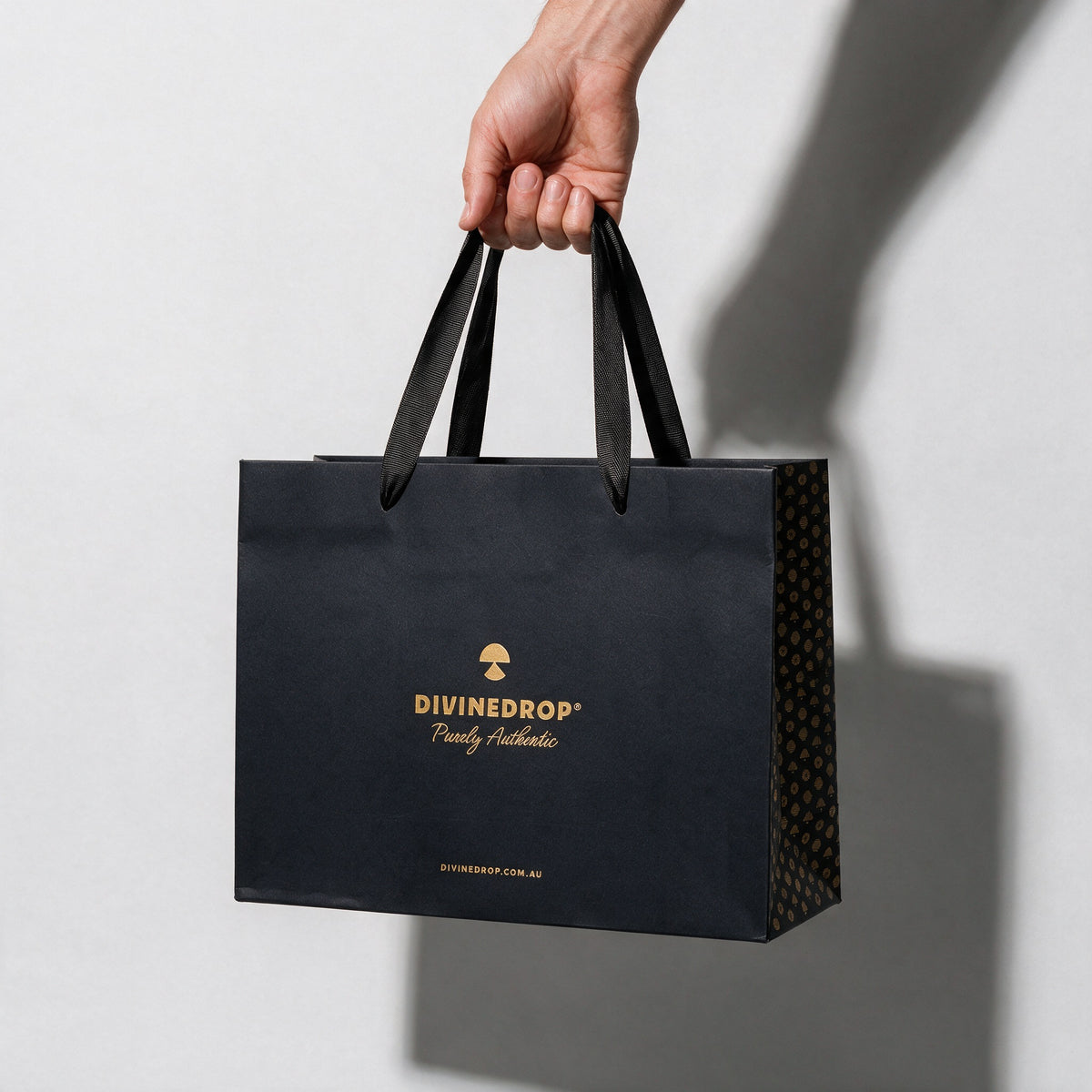 Hand carrying a black DIVINEDROP gift bag by its ribbon handles, on a light grey background