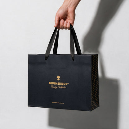 Hand carrying a black DIVINEDROP gift bag by its ribbon handles, on a light grey background
