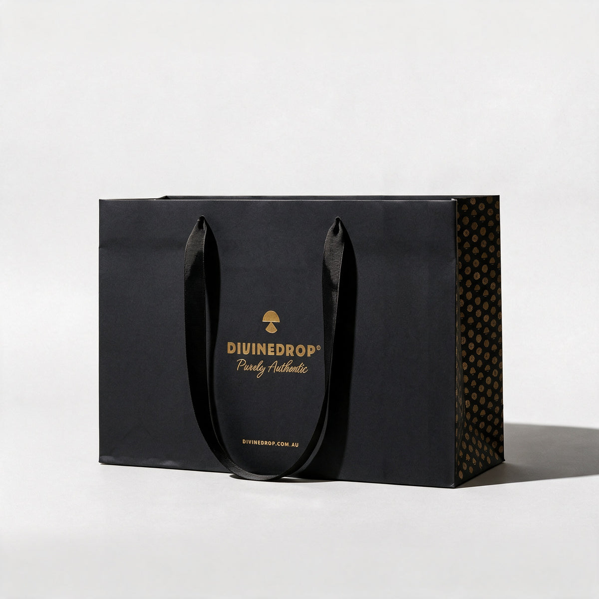 Black DIVINEDROP gift bag with gold foil logo, black ribbon handles and gold pattern side panel, on a light grey background