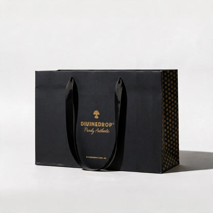 Black DIVINEDROP gift bag with gold foil logo, black ribbon handles and gold pattern side panel, on a light grey background