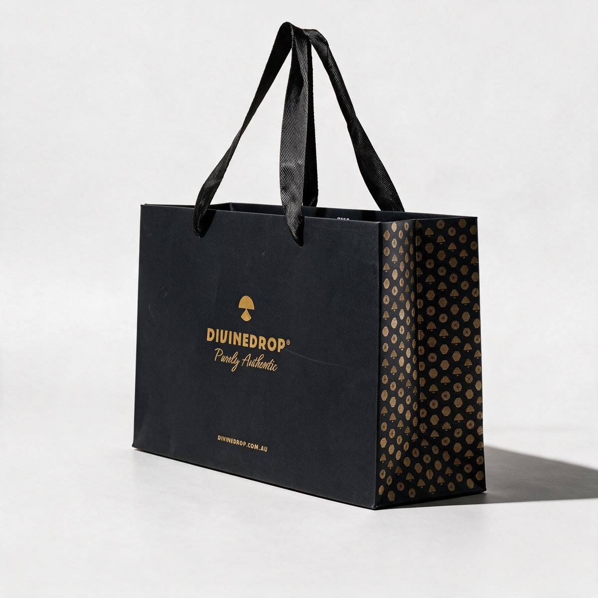 DIVINEDROP black gift bag seen from the side, showing the gold pattern side panel and black ribbon handles, on a light grey background