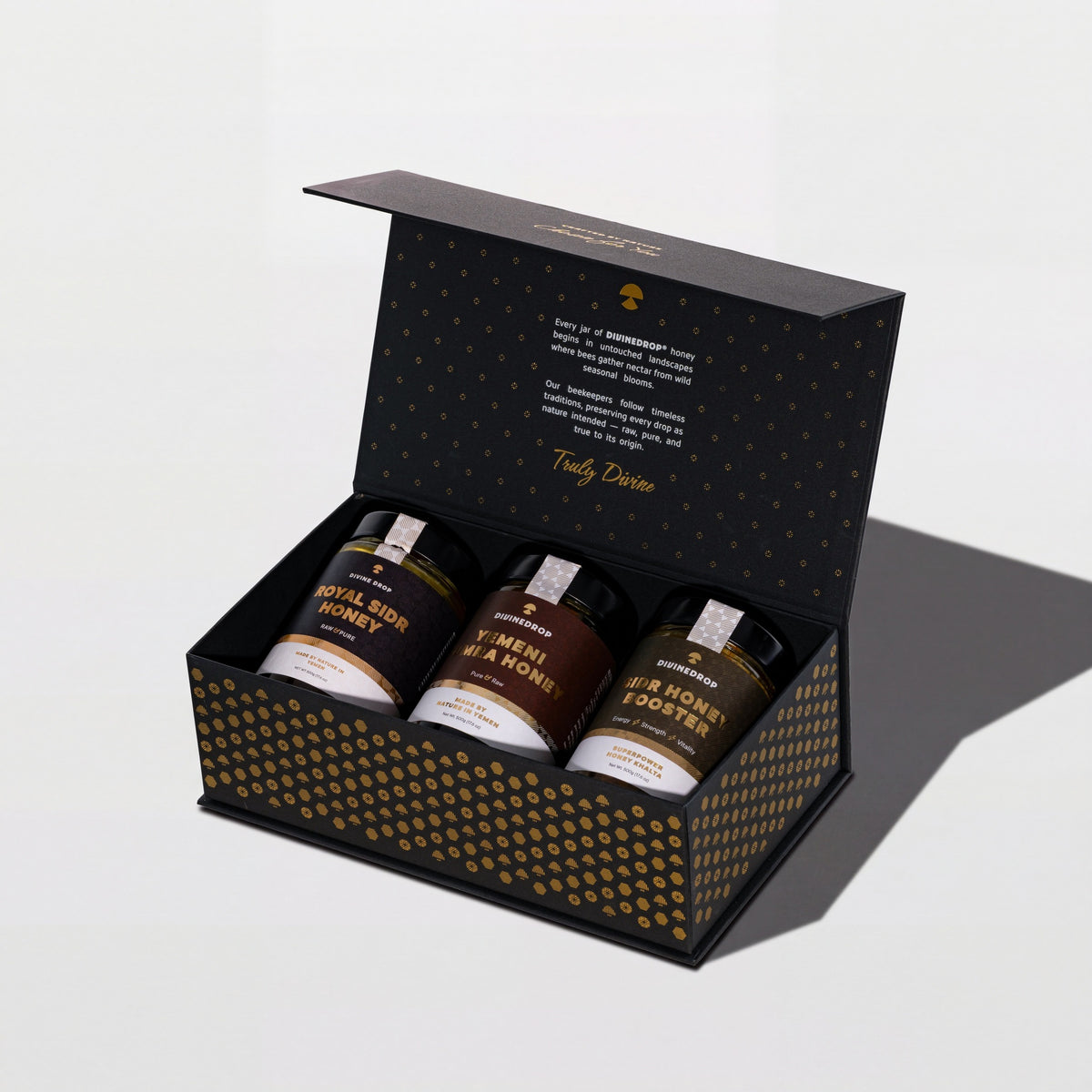 DIVINEDROP black gift box with gold pattern, open, holding three jars of Yemeni honey, on a light grey background