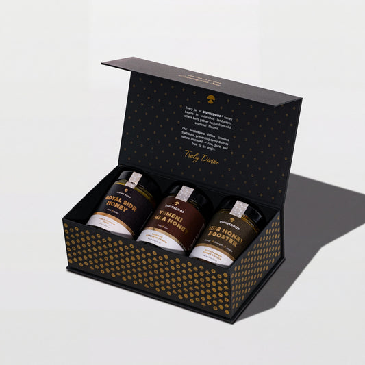DIVINEDROP black gift box with gold pattern, open, holding three jars of Yemeni honey, on a light grey background